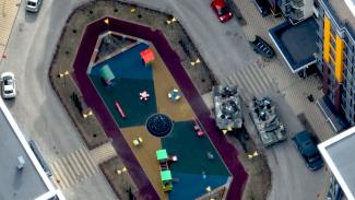 Still from One Day in Ukraine. Aerial view image of a children's playground, set in the middle of a roundabout. Beside the playground are parked cars, and two military tanks.