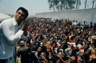 Still from When We Were Kings. Muhammad Ali is leaning out of the window of a bus, to a crowd of people waving and cheering.