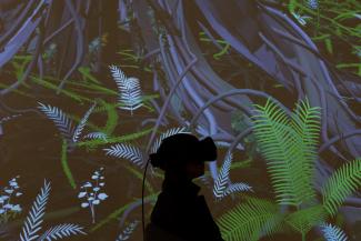 Silhouette of a person wearing a VR headset, stood in front of a projection of digitally rendered trees and leaves.