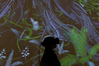 Silhouette of a person wearing a VR headset, stood in front of a projection of digitally rendered trees and leaves.