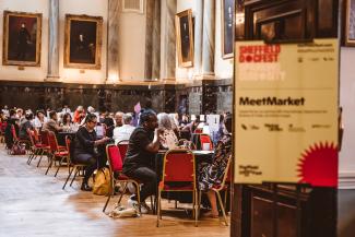 A hall filled with round tables, and people taking meetings around them. To the right of the image, slighty blurred and out of focus is a sign which reads 'MeetMarket'
