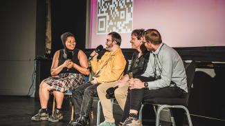 Four people sat on stage in a row, in front of a large cinema screen. Two are holding microphones towards them and are smiling.