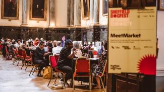 A hall filled with round tables, and people taking meetings around them. To the right of the image, slighty blurred and out of focus is a sign which reads 'MeetMarket'