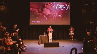 A person with dark hair, wearing a long red shirt is stood in front of a podium, facing an audience of people who are sat in stalls at the edges of the room. On a screen behind the podium is a film still showing two people kissing. Over the image in pink text reads: Anhell69.