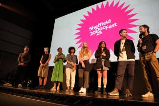 A group of filmmakers stand on a stage looking to the person on the far right who is holding a microphone. Behind them, a screen shows the words 'Sheffield DocFest 2023'