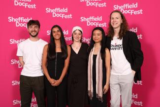 Five young people stand in a line with wide smiles on their faces. Being them is a hot pink step and repeat board with the Sheffield DocFest logo (the words Sheffield DocFest in white) repeatedly pointed on it.