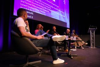 Panel of people sat on chairs on stage with a woman stood at a lectern facing away from the camera in the foreground