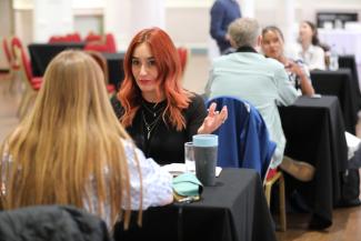 A line of tables with people either side of each table chatting animatedly. The most prominent person in the image is a person in the middle of the photo with long red hair. 