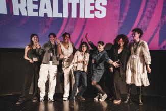 A group of people stood on a stage looking joyful, with smiles on their faces and their hands in the air.