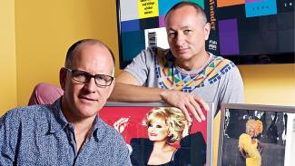 Portrait of Fenton Bailey, pictured right, and Randy Barbato, pictured left, sitting at a desk with two computer screens against a yellow wall.