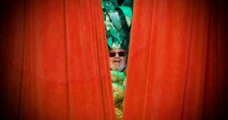 Large red theatre curtains cover most of the image, except for the very center where a man's face peeks through. The man is wearing a pineapple costume and a pair of large red sunglasses.