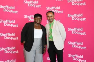 Grenfell: Uncovered director Olaide Sadiq, pictured with Festival Creative Director Raul Niño Zambrano smile as they stand against a bright pink and white branded backdropat Sheffield DocFest 2025
