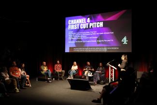 Panel of people sat on chairs on stage with a woman stood at a lectern facing away from the camera on the right.