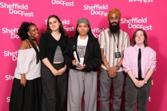 Five young people stand in a line with wide smiles on their faces. Being them is a hot pink step and repeat board with the Sheffield DocFest logo (the words Sheffield DocFest in white) repeatedly pointed on it.